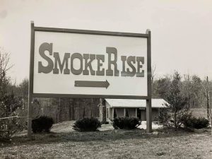 Black and white photograph of the original wooden Smoke Rise entrance sign, with an arrow pointing toward the subdivision entrance